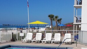 A pool area with lounge chairs, a flag, and a view of the beach. - Ambassador 128- Gulf Front - Pet Friendly (Panama City Beach)