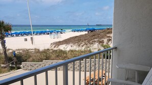 A balcony with a view of a beach, featuring a flag, beach umbrellas, and a pier in the distance. - Ambassador 128- Gulf Front - Pet Friendly (Panama City Beach)