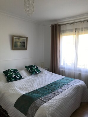 Een opgemaakt bed met een sprei, twee kussens, een ingelijste foto aan de muur en een raam met gordijnen.