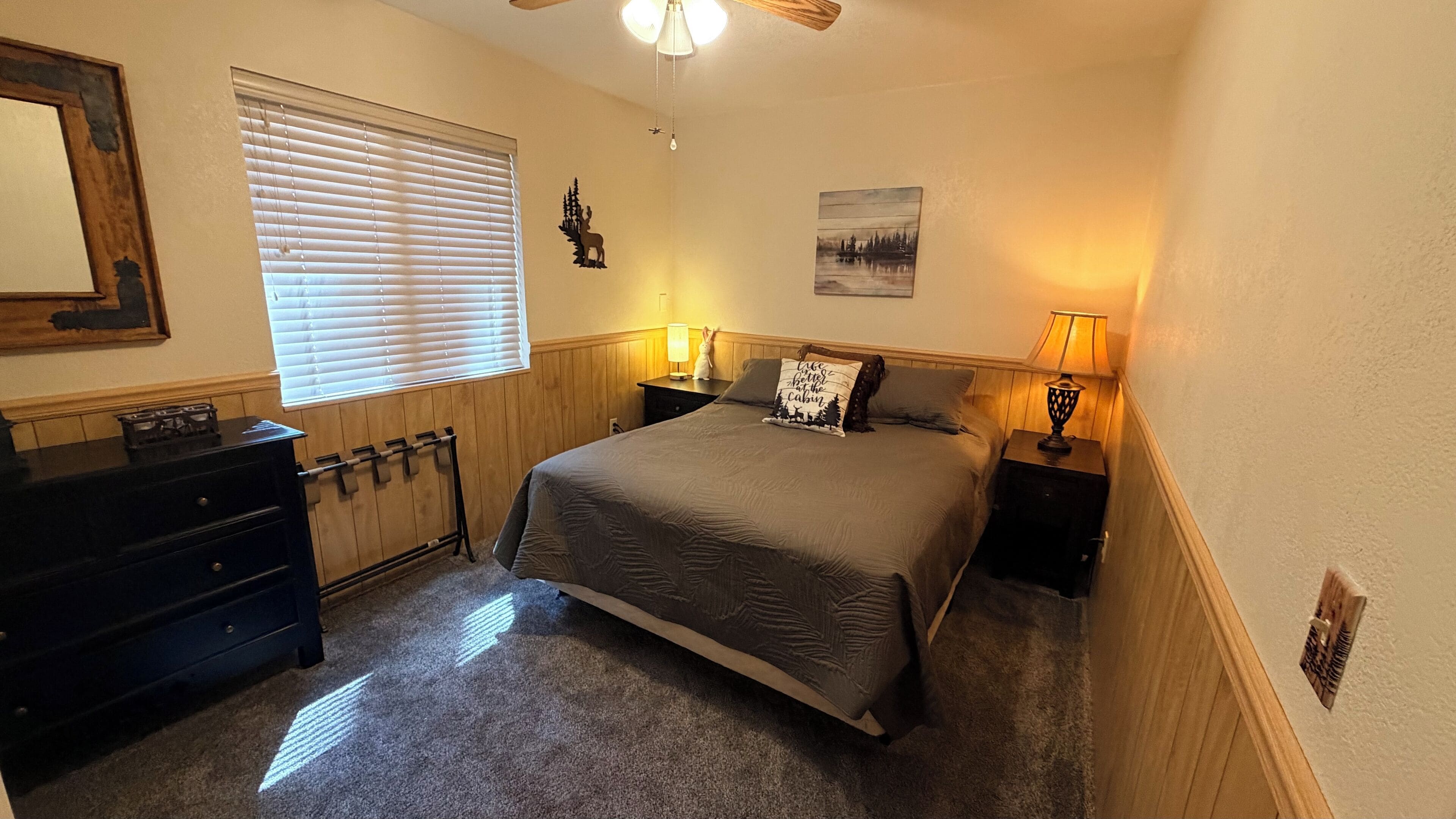 A bedroom with a bed, two bedside tables with lamps, a dresser, a window with blinds, a ceiling fan, and a wall-mounted picture.