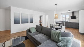 A modern apartment with a grey sofa, a glass coffee table, and a dining area with a table and chairs. There is a kitchen with grey cabinets and a hanging light fixture. - Awesome apartment in Lörrach with WiFi (Lörrach)