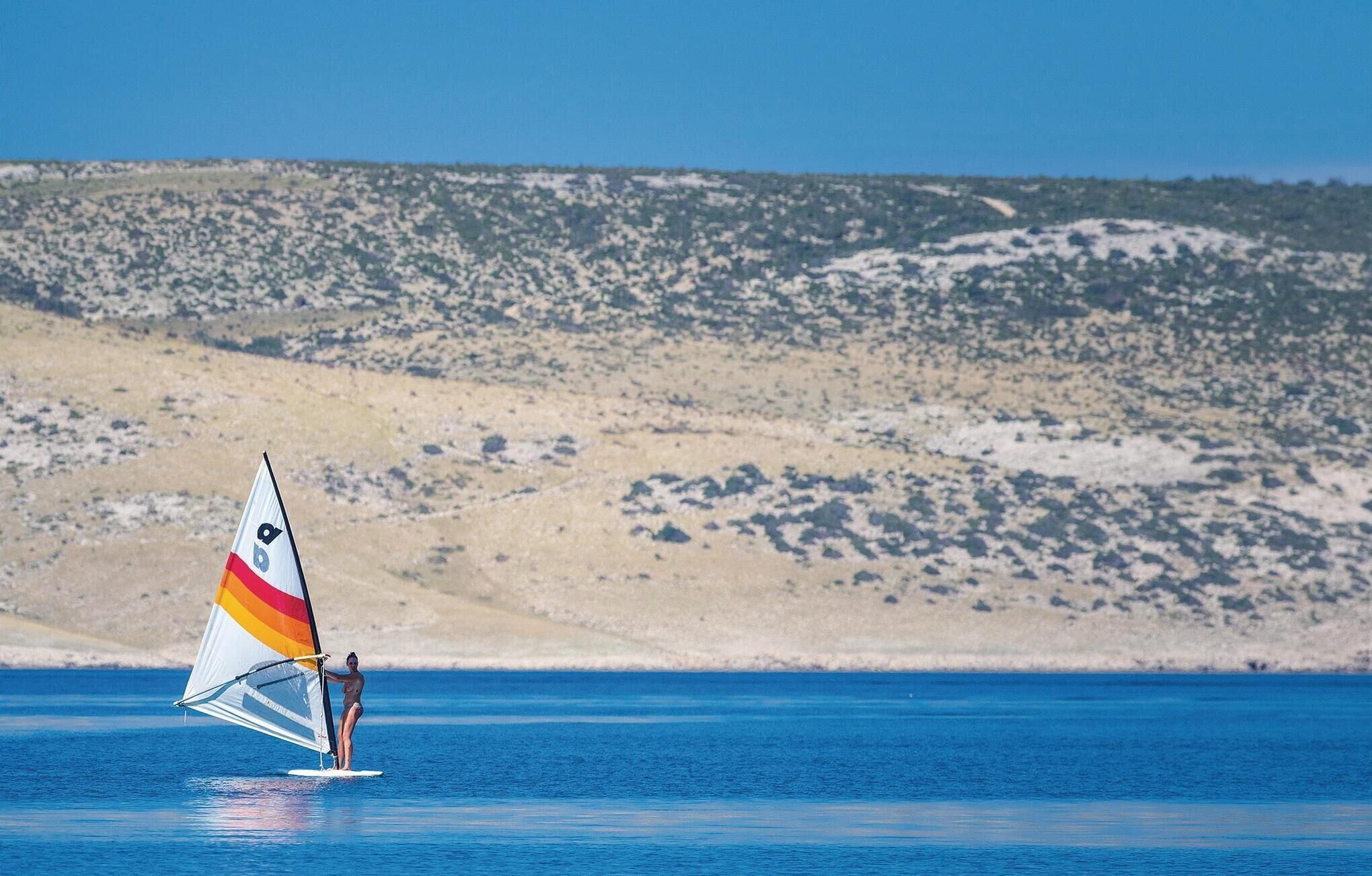 Une personne pratiquant la planche à voile par une journée ensoleillée, avec une plage de sable et un paysage vallonné en arrière-plan.