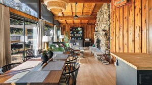 A modern interior with a wooden ceiling, stone wall, and a fireplace. There is a dining area with a table and chairs, and a kitchen area with a stove and cabinets. - Family Cabin, Dog, Ht Tb, 15 min Yosemite, Valentine's Day + Firefall Packages (Oakhurst)