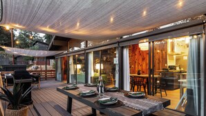 An outdoor dining area with a wooden deck, a long table set for a meal, and a view into the kitchen through glass doors. - Family Cabin, Dog, Ht Tb, 15 min Yosemite, Valentine's Day + Firefall Packages (Oakhurst)