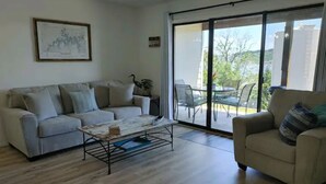 Un soggiorno con divano, poltrona e tavolino. Una porta vetrata scorrevole dà accesso a una zona esterna con sedie.