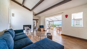 Un salon spacieux avec un grand canapé bleu, une télévision et une cheminée. Il y a un balcon avec des sièges d’extérieur et une vue sur le paysage.