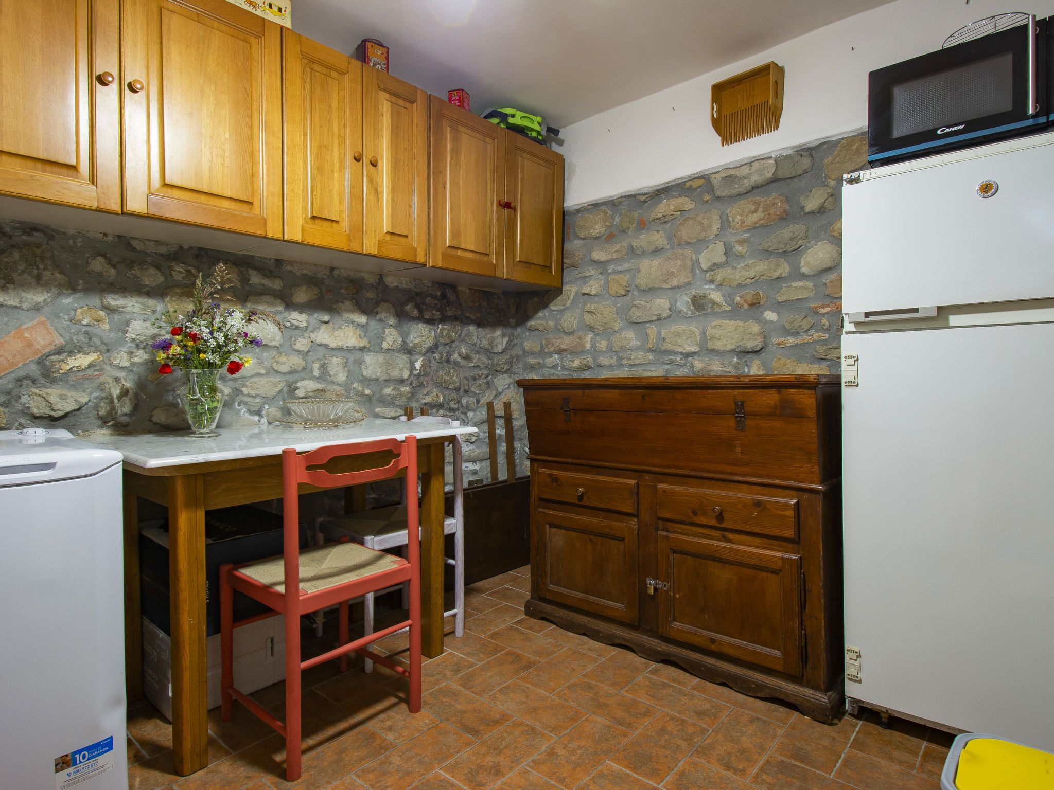A kitchen with wooden cabinets, a stone wall, a white refrigerator, and a red chair.