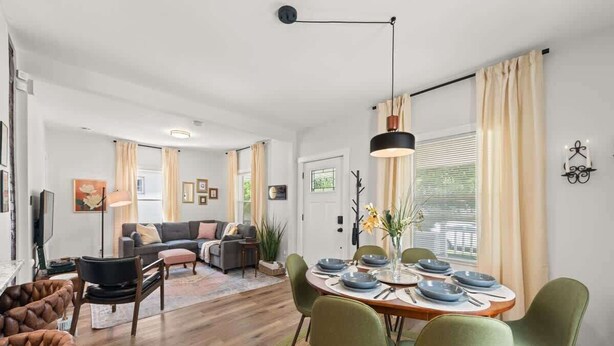 Ein Essbereich mit einem Holztisch, gedeckt für vier Personen, umgeben von grünen Stühlen. Im Hintergrund befindet sich eine Couch und ein Couchtisch, darüber hängt eine Lampe.
