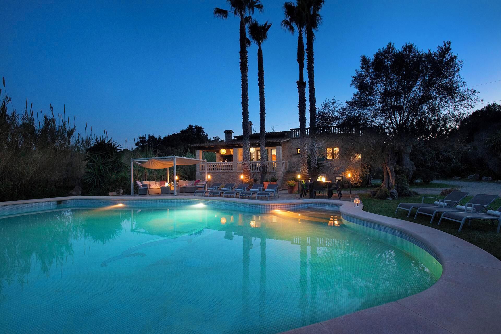 A swimming pool with lounge chairs and a covered seating area, surrounded by tall palm trees and a stone building with outdoor lighting.