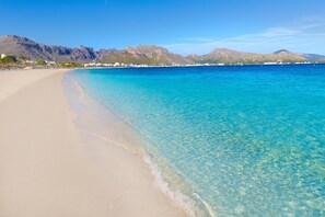 Une plage à l’eau turquoise et limpide, un rivage sablonneux et des montagnes en arrière-plan.