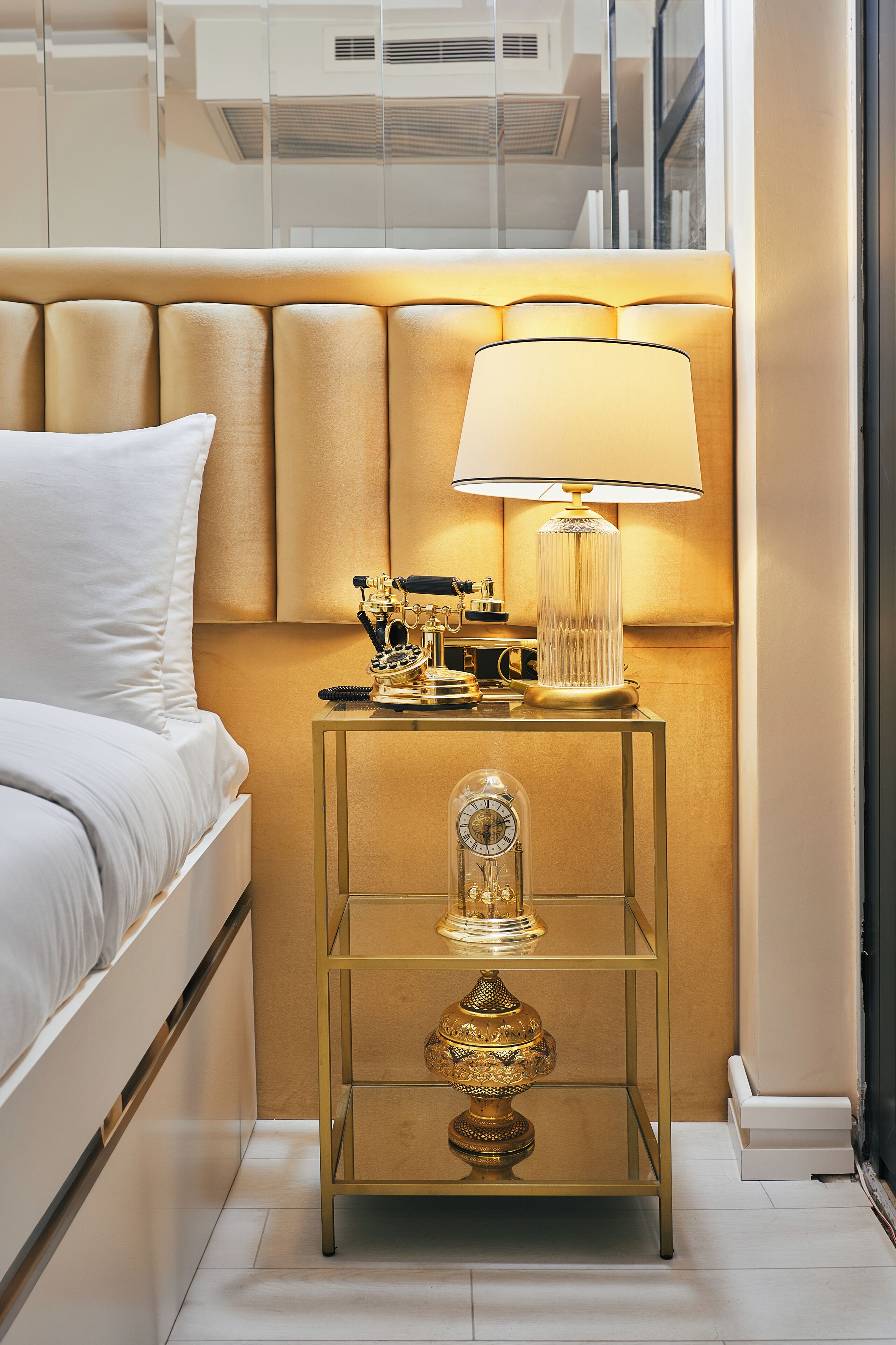 A modern hotel room with a neatly made bed, a bedside lamp, a small table with decorative items, and a glass cabinet.