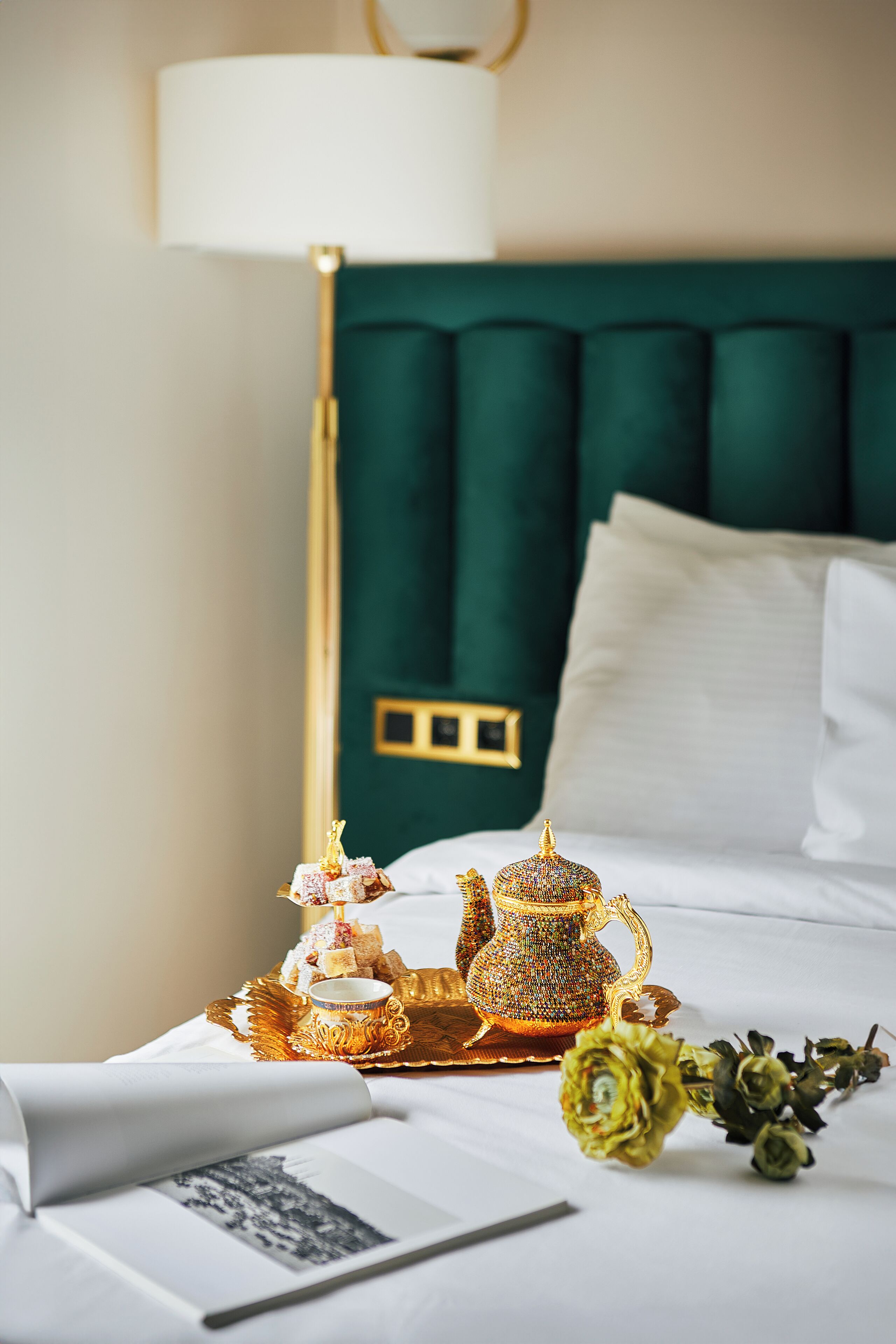 Una cama bien hecha con un cabecero tapizado en verde, una mesita de noche con lámpara y una bandeja con tetera y bombones.