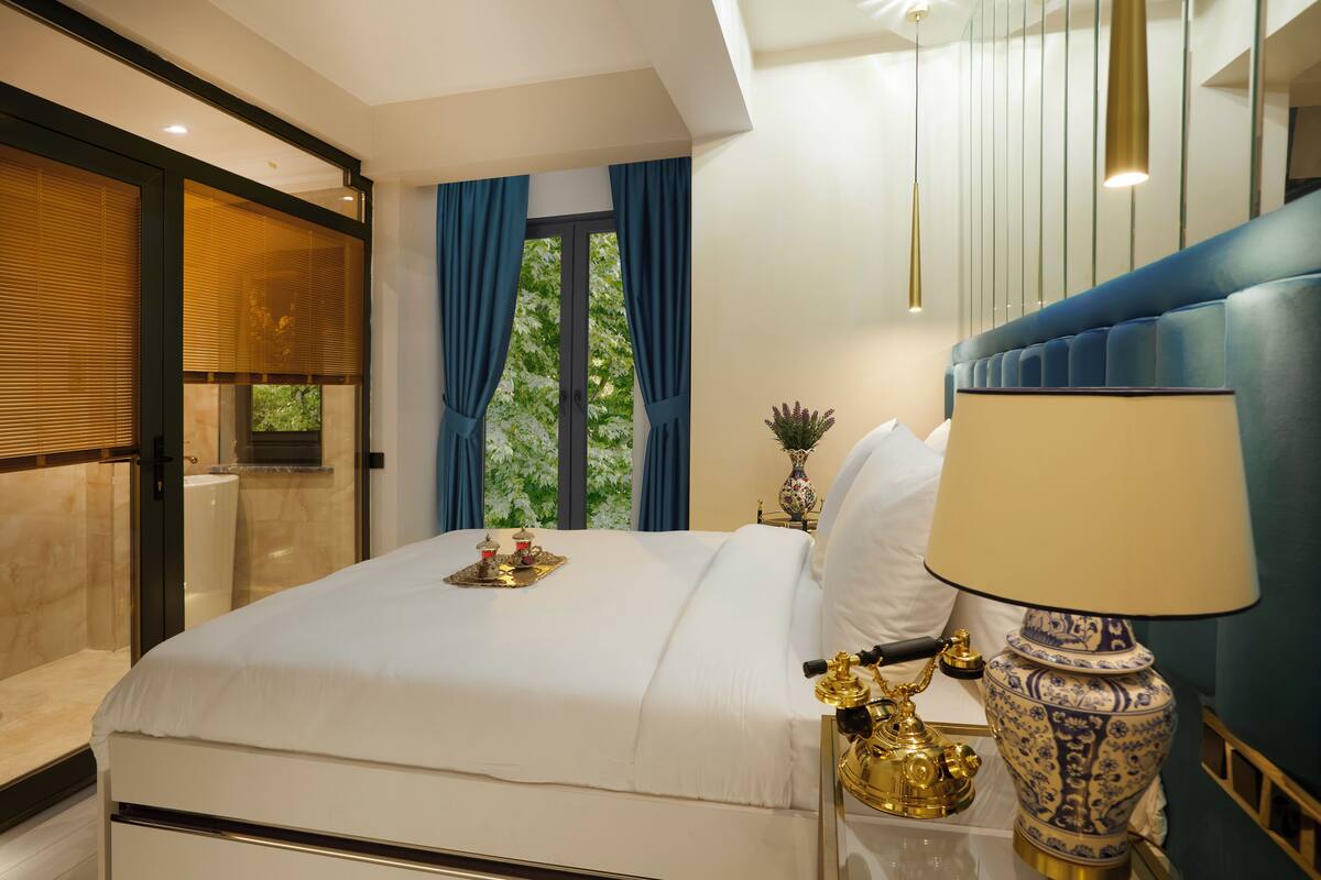 Quarto de hotel moderno com uma cama grande, mesa de cabeceira com abajur e vista para o verde pela janela.