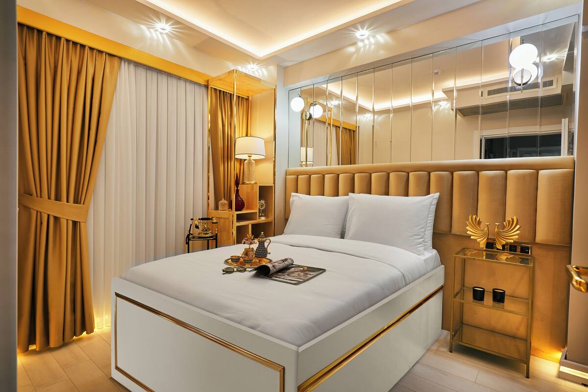 Quarto de hotel moderno com uma cama grande, mesa de cabeceira com abajur, espelho e uma estante decorativa.