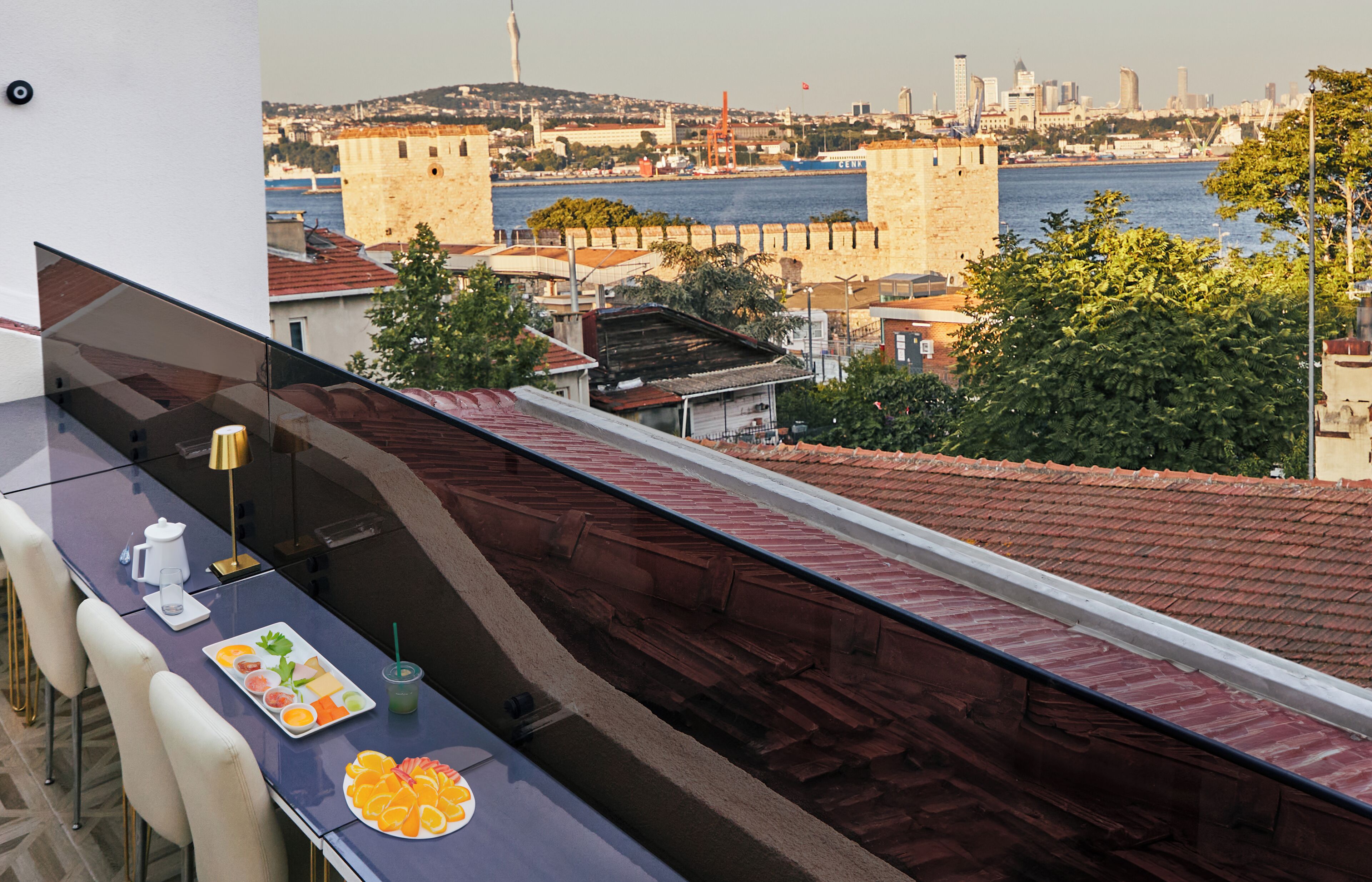 Un balcon avec une table dressée pour deux personnes, offrant une vue sur un paysage urbain avec des bâtiments historiques et une étendue d’eau.