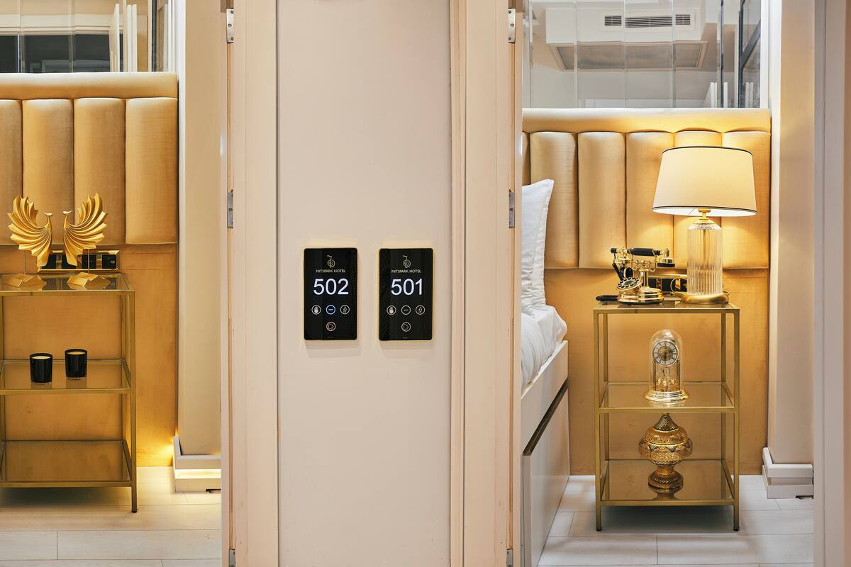 Quarto de hotel com cama, mesas de cabeceira, abajur e um objeto decorativo. Os números 502 e 501 estão exibidos na parede.