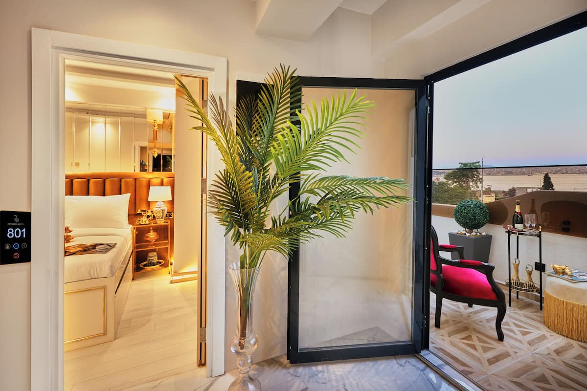 Quarto de hotel moderno com cama, mesa de cabeceira, abajur e uma janela grande com vista para a paisagem urbana.