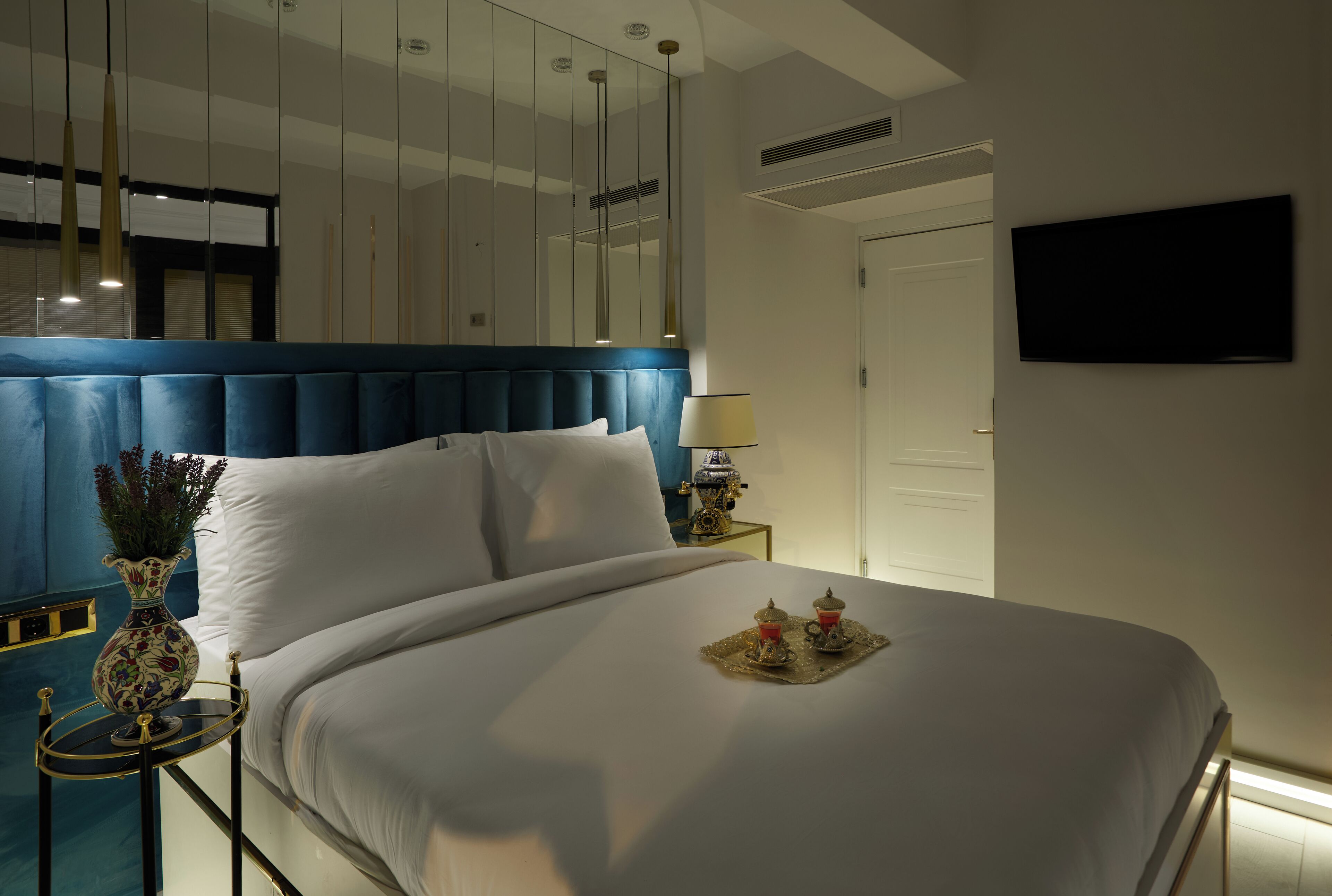 Una cama bien hecha con sábanas blancas, cabecera azul, mesita de noche con lámpara y un jarrón con flores.