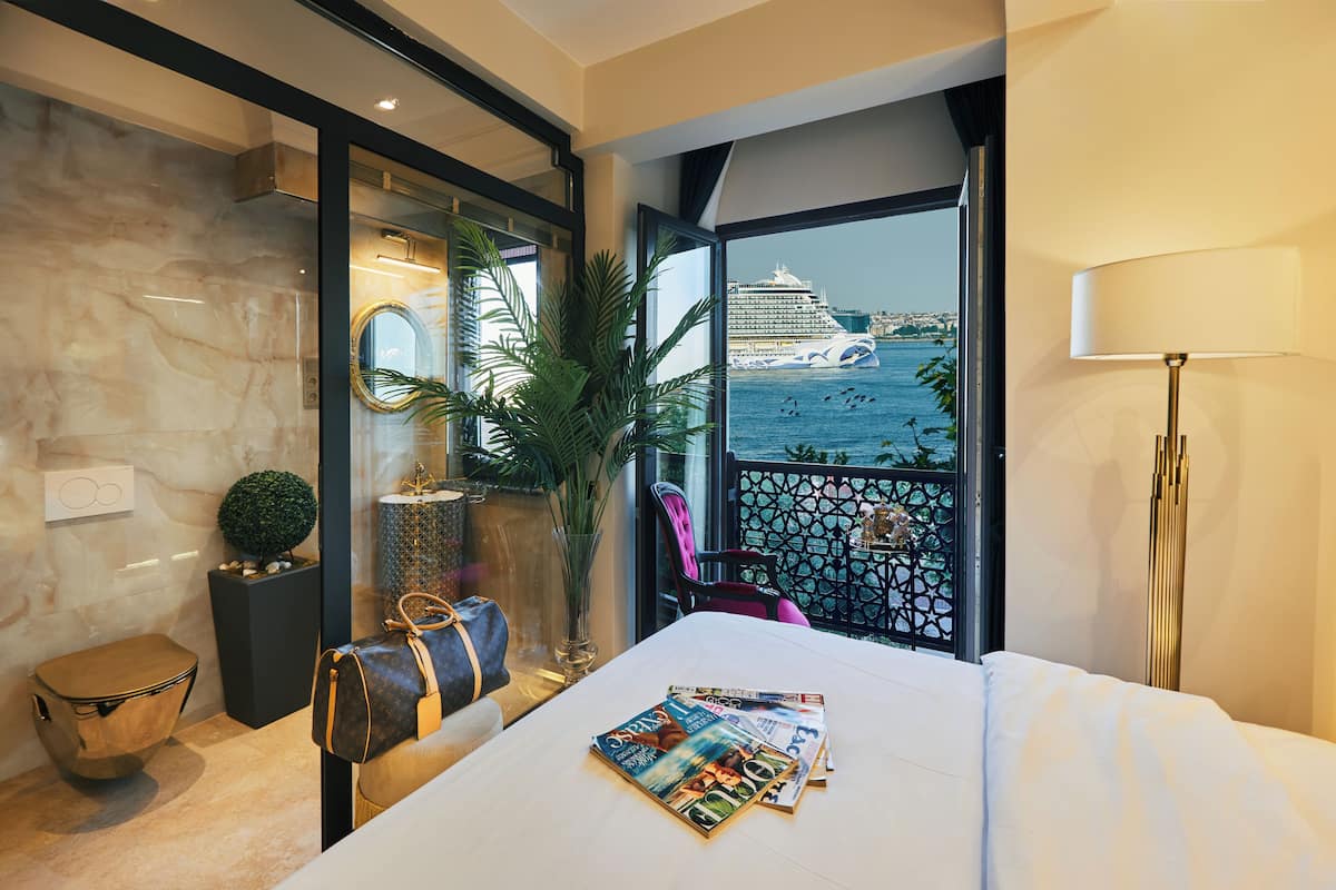 Quarto de hotel com uma cama grande, vista para um navio de cruzeiro, varanda com guarda-corpo e um abajur.