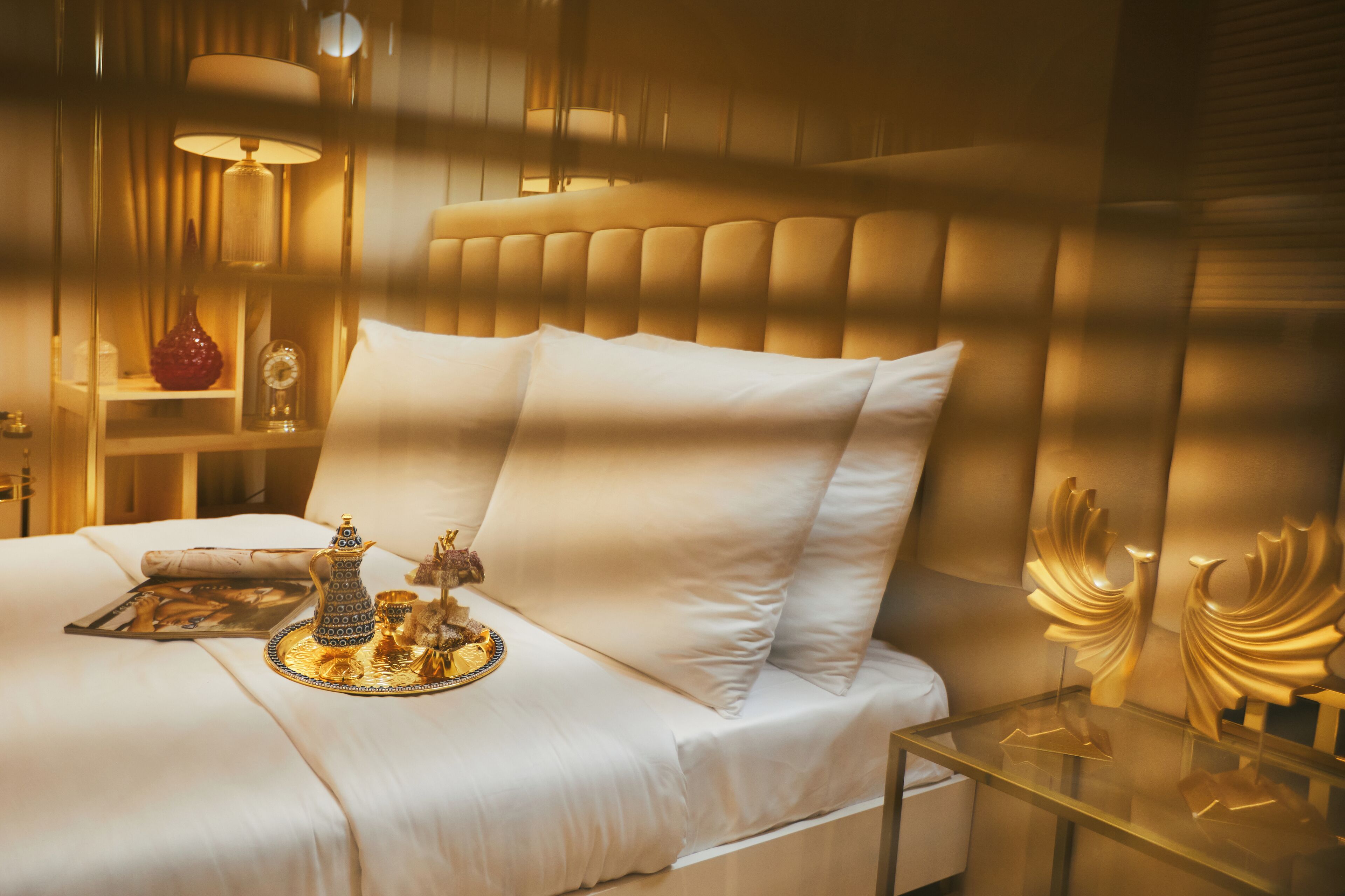 A neatly made bed with white linens, a bedside table with a lamp, and decorative items including a tray with a teapot and sugar bowl.