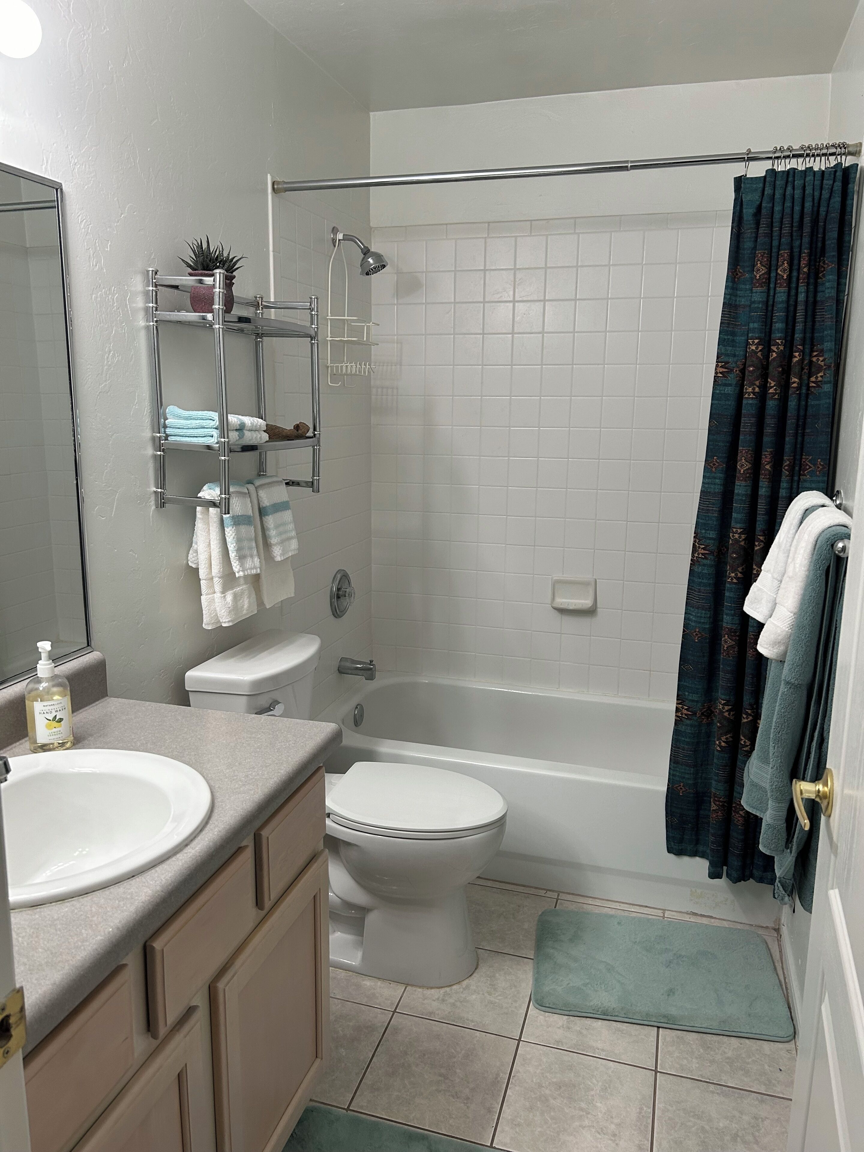 A bathroom with a white sink, toilet, and bathtub. There is a towel rack with towels, a small shelf with a plant, and a shower curtain.