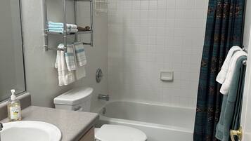 A bathroom with a white sink, toilet, and bathtub. There is a towel rack with towels, a small shelf with a plant, and a shower curtain.
