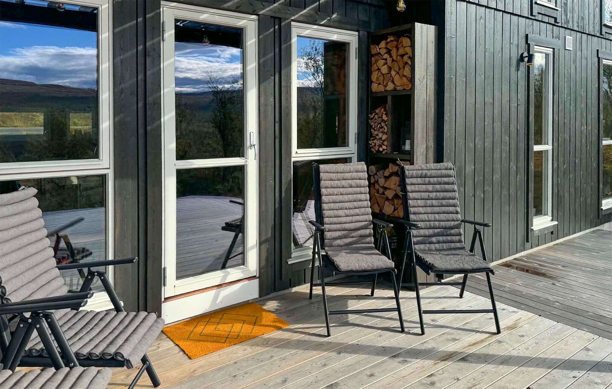 Een terras met twee stoelen en een deurmat, een houten muur en een raam met uitzicht op de lucht en bomen.