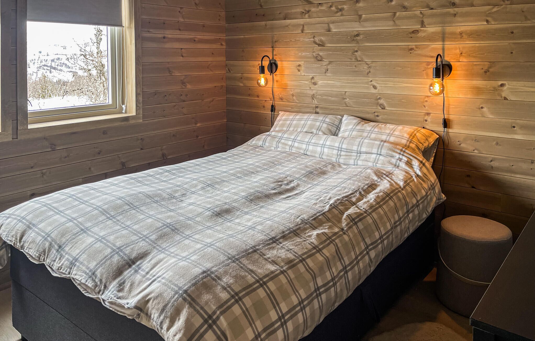 A bedroom with a bed, two wall-mounted light fixtures, a window, and a small round stool.