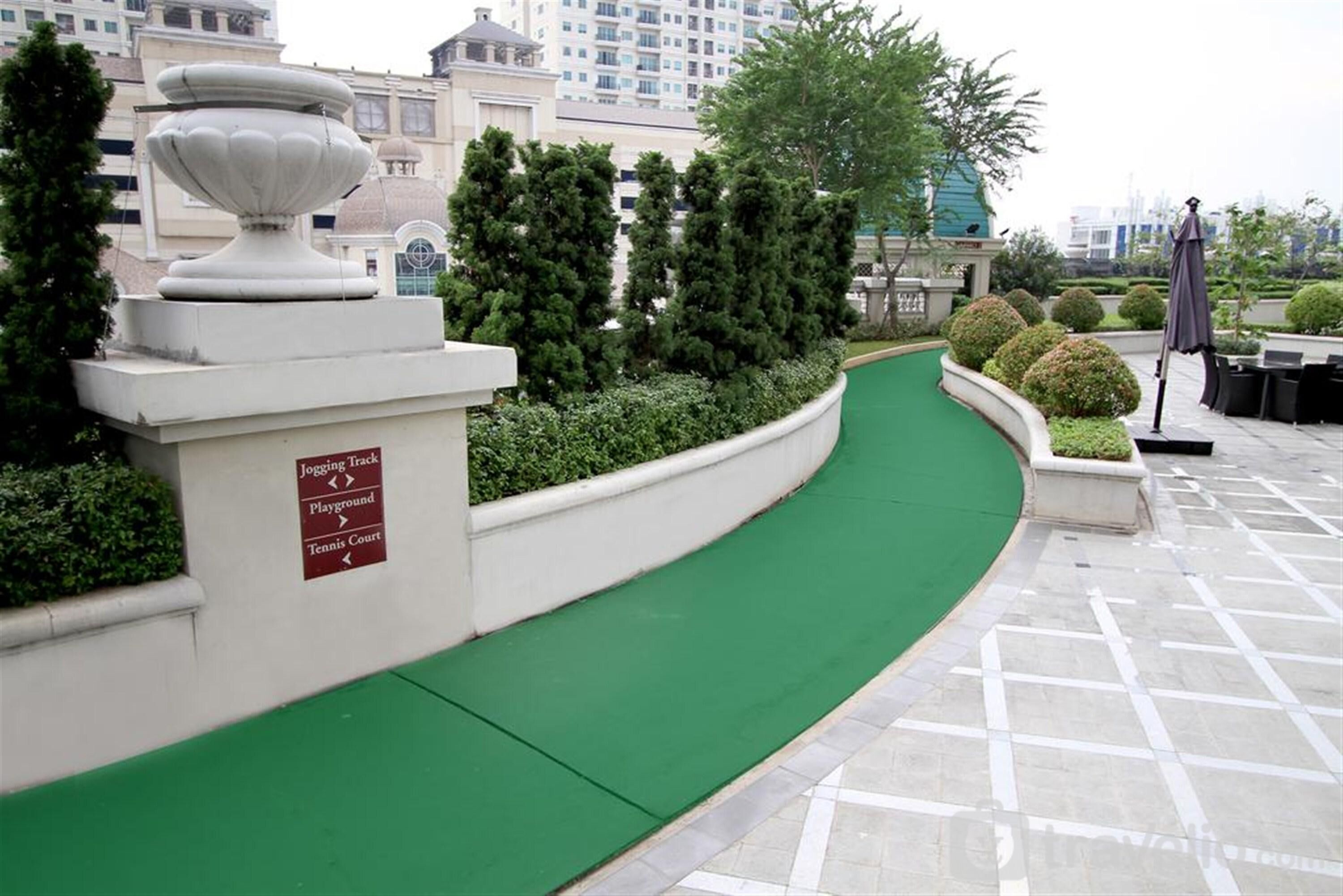 A curved green pathway leading to a tennis court, surrounded by neatly trimmed bushes and a decorative urn.
