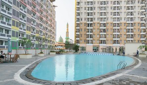 A swimming pool surrounded by multi-story residential buildings with balconies, a mosque with a green dome, and outdoor seating areas.