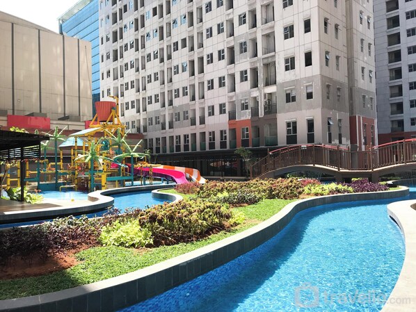 A swimming pool with a water slide, surrounded by greenery and a playground.
