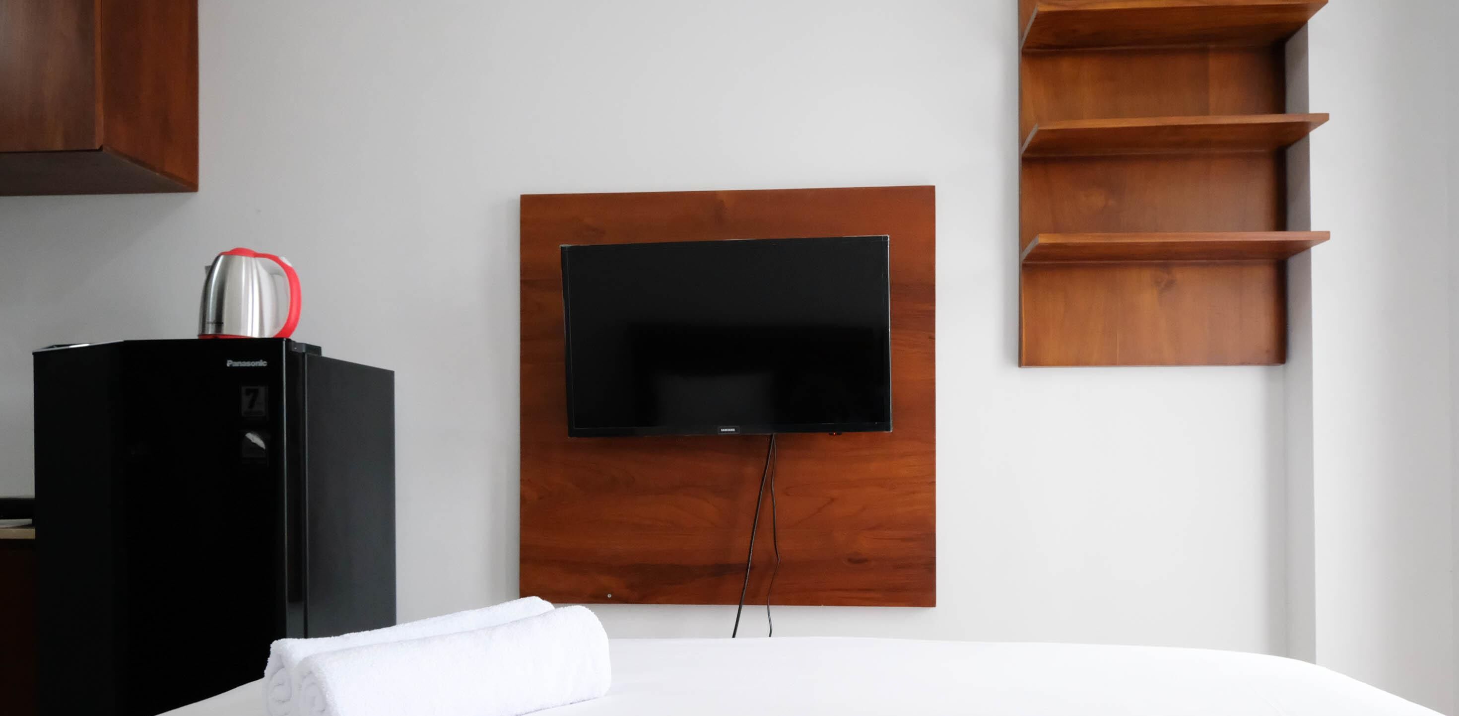 Una habitación de hotel con una cama, un televisor de pantalla plana montado en un panel de madera, un minibár negro y una estantería de madera.