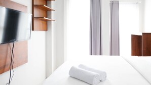 Habitación de hotel con televisión de pantalla plana montada en la pared, estantes de madera, cama con ropa blanca y ventana con persianas verticales.