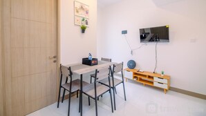 A room with a wooden door, a dining table with four chairs, a TV mounted on the wall, and a small wooden shelf with a clock and a plant. - Comfort And Nice 2Br At Tokyo Riverside Pik 2 Apartment (Teluknaga)
