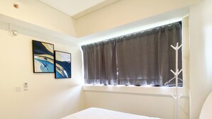 A hotel room with a bed, a window with curtains, a coat rack, and two paintings on the wall. - Good Deal And Modern Look 3Br Meikarta Apartment (Cikarang)