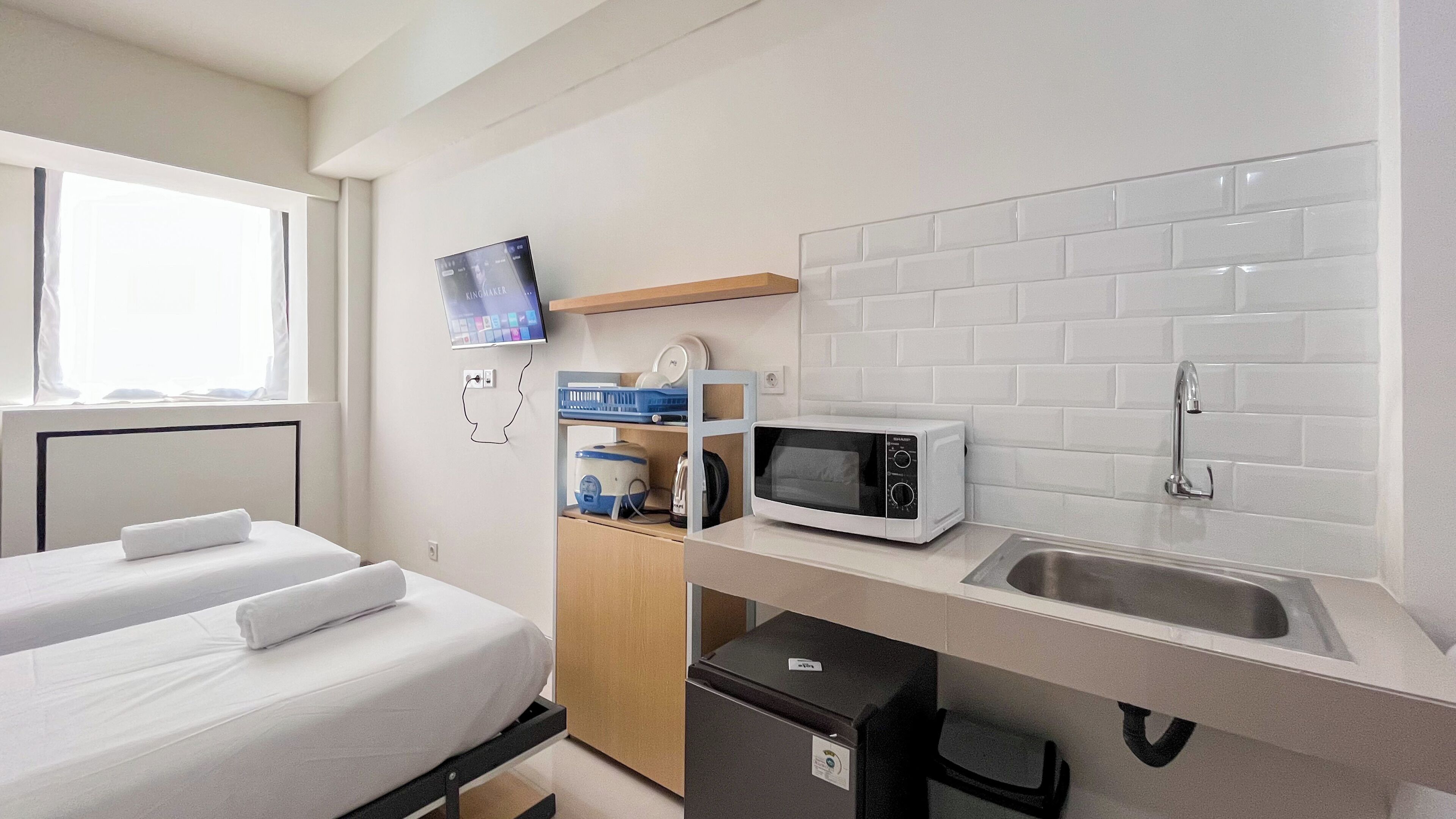 A compact hotel room with a bed, a small kitchenette, a microwave, a sink, and a television mounted on the wall.