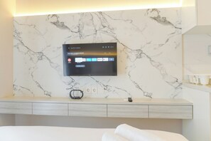 A modern hotel room with a marble wall, a mounted flat-screen TV, a bedside table with a remote control, and a bed with a white comforter. - Good And Modern Studio At Sky House Alam Sutera Apartment (Tangerang)