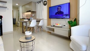 Una sala moderna con un televisor de pantalla plana montado en la pared, un comedor con sillas blancas y una pequeña mesa redonda con una cesta tejida.