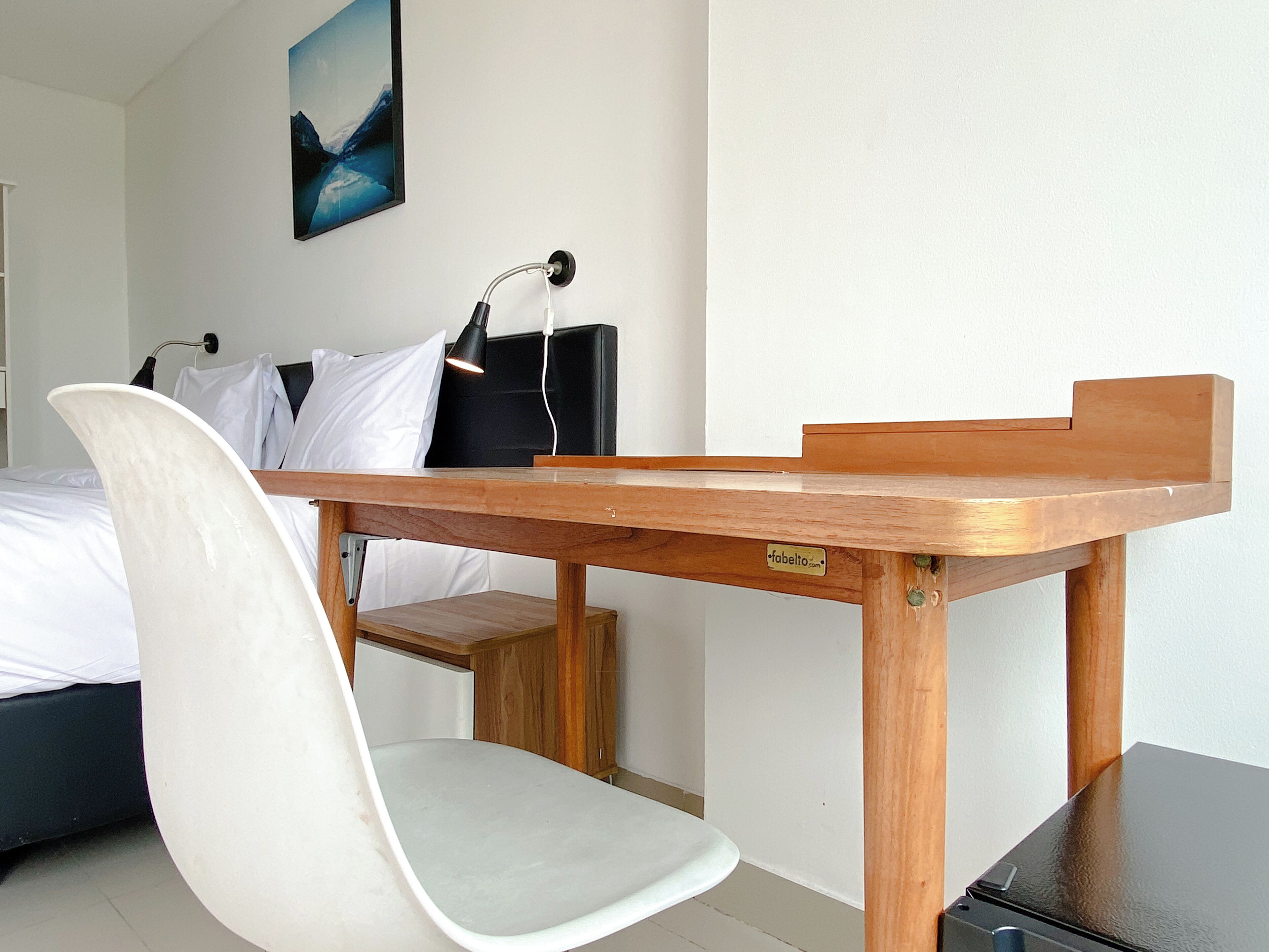 Ein Hotelzimmer mit einem Holztisch, einem weißen Stuhl, einem Bett mit weißer Bettwäsche, einer schwarzen Lampe und einem Gemälde an der Wand.