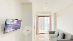 A modern living room with a flat-screen TV mounted on the wall, a standing fan, and a grey sofa. - Best Choice And Comfy 2Br Daan Mogot City Apartment (Jakarta)