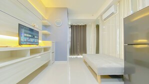 A modern hotel room with a flat-screen TV on a white shelf, a bed, a refrigerator, and a window with curtains. - Brand New And Modern 2Br At Daan Mogot City Apartment (Jakarta)