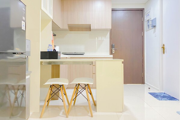 A compact kitchen with a refrigerator, a small countertop, two stools, and a door in the background.