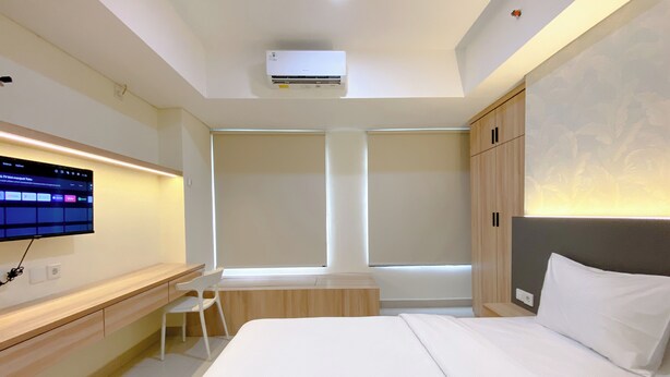 A modern hotel room with a large bed, a desk, a TV mounted on the wall, and a window with blinds. - Simply Studio Pollux Chadstone Apartment (Cikarang)