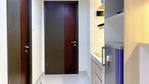 A hallway with a closed door, a refrigerator, and a shelf with items. - Simply Studio Pollux Chadstone Apartment (Cikarang)
