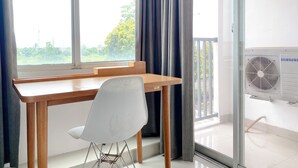 A room with a wooden desk, a white chair, a large window with curtains, and a Samsung air conditioner.