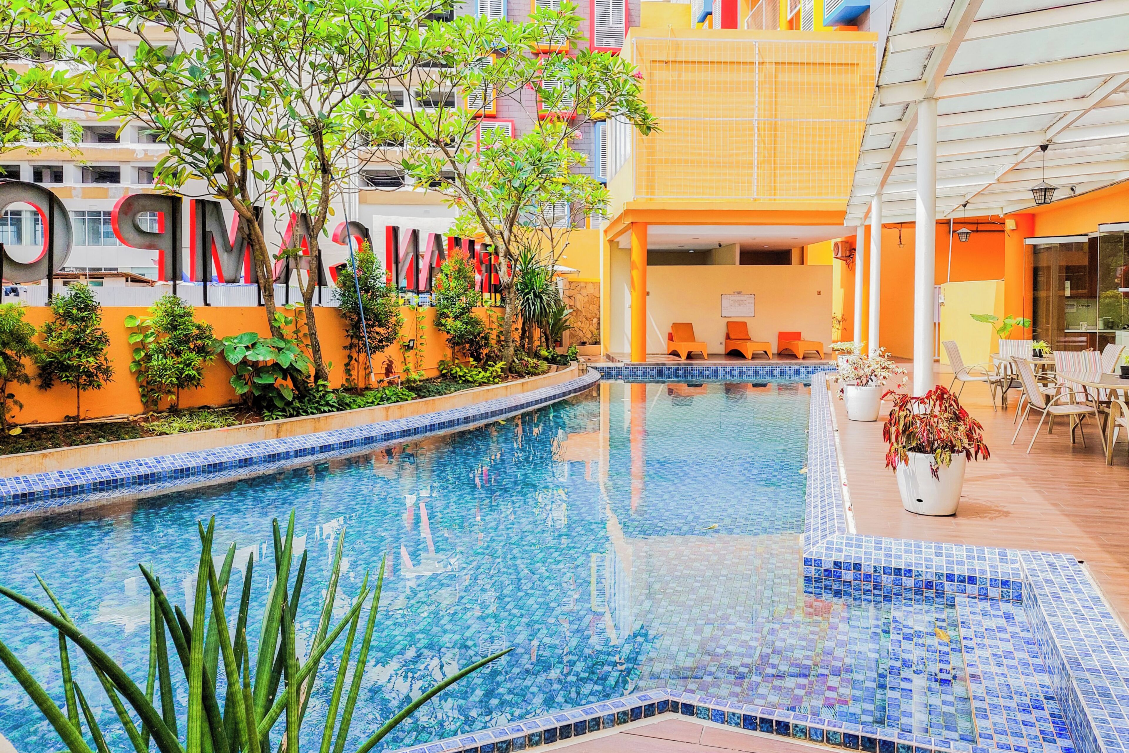 A swimming pool with a tiled edge, surrounded by plants and a building with a colorful facade.