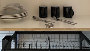 Uma prateleira na cozinha com pratos empilhados, canecas pretas e utensílios.