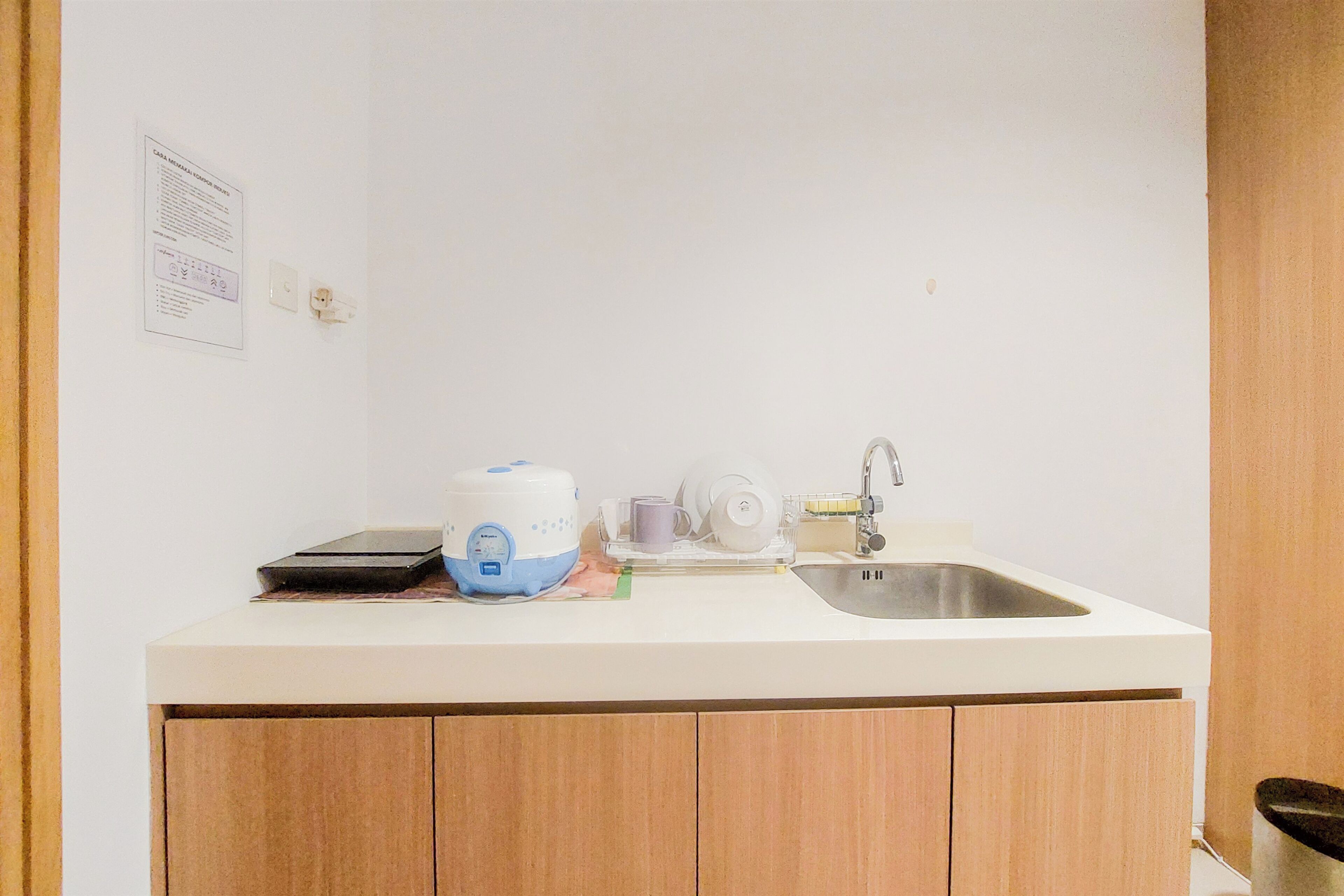 Una pequeña cocina con encimera blanca, fregadero y armarios de madera. Se observa una cocedora de arroz, un escurreplatos y algunos platos sobre la encimera.