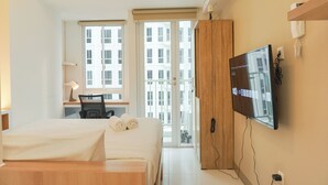 A modern hotel room with a bed, desk, chair, TV, and large windows offering a city view. - Comfy And Best Deal Studio At Tokyo Riverside Pik 2 Apartment (Teluknaga)