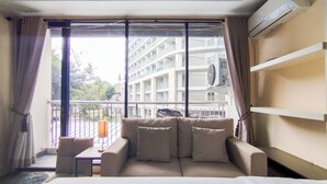 Et hotelværelse med seng, sofa, et lille bord og en balkon med udsigt til en bygning og træer.